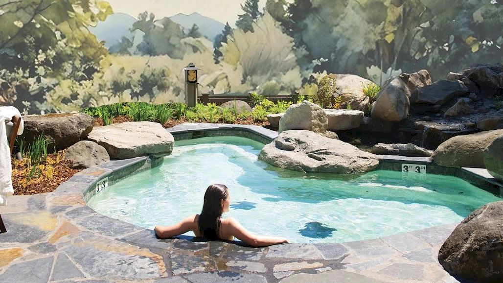 A person relaxes in a rocky hot tub with scenic forest views surrounding the area, creating a tranquil outdoor retreat.