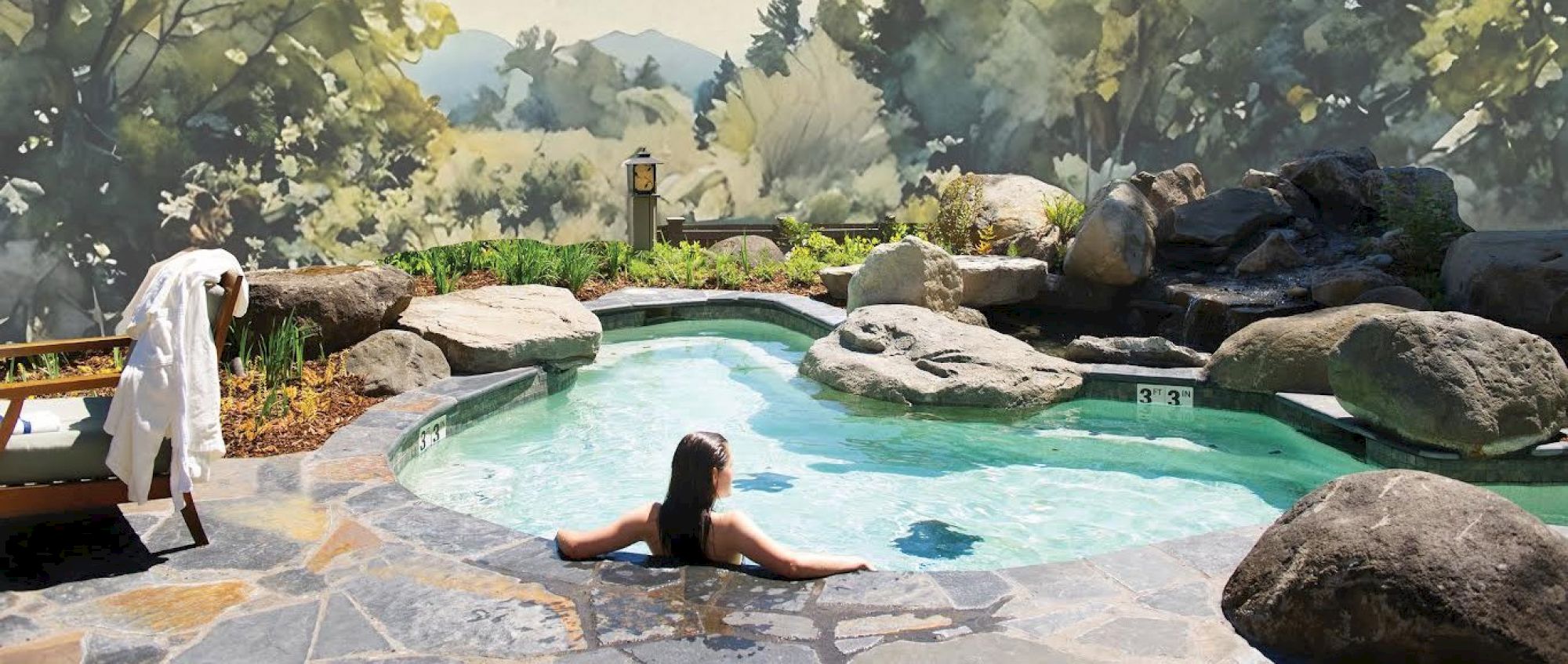 A person relaxes in a rocky hot tub with scenic forest views surrounding the area, creating a tranquil outdoor retreat.