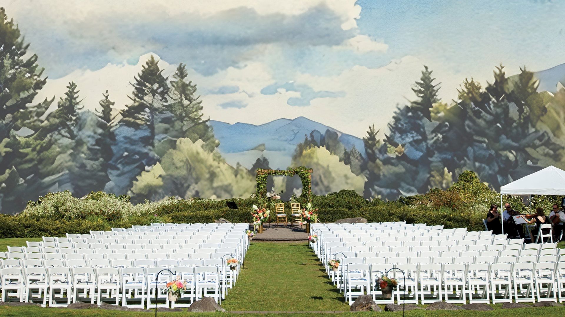 Outdoor wedding setup with white chairs facing an altar under an arch, surrounded by greenery and mountains, and a tent on the right.