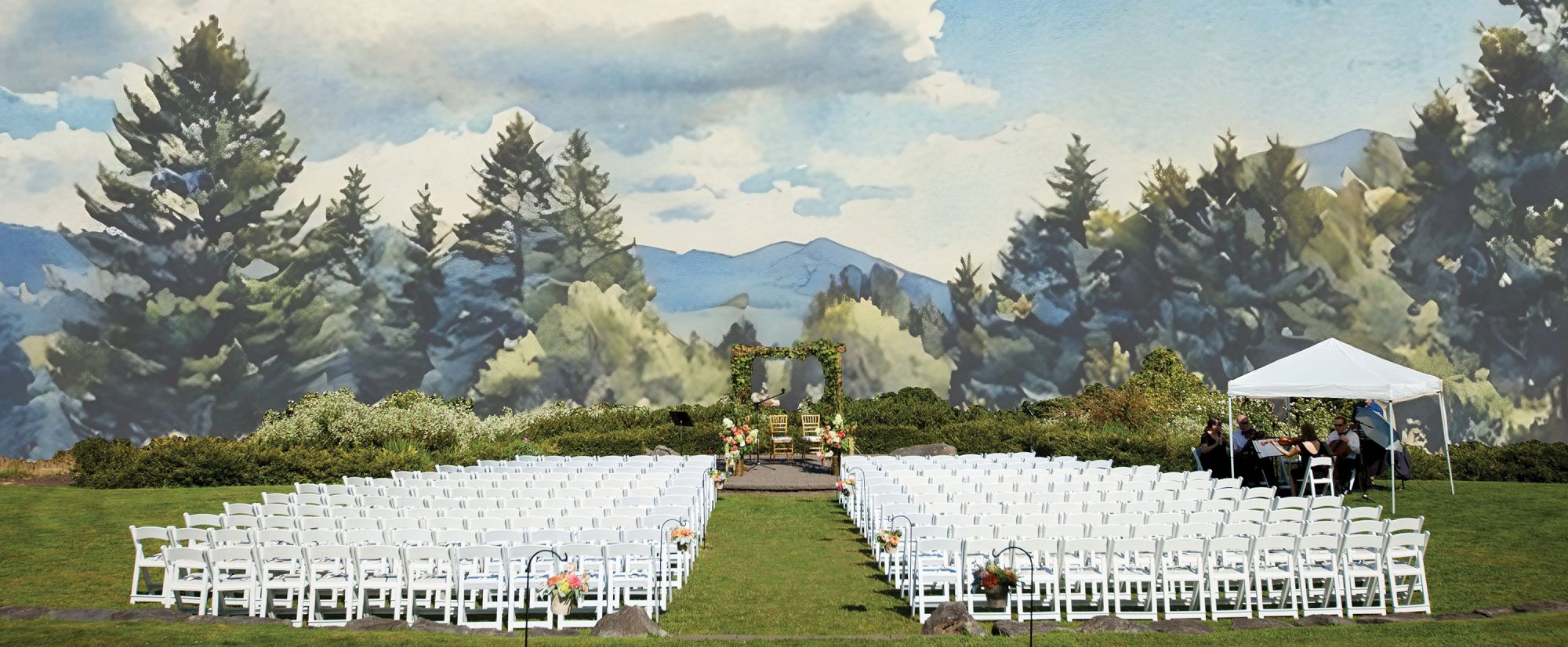 Outdoor wedding setup with white chairs facing an altar under an arch, surrounded by greenery and mountains, and a tent on the right.