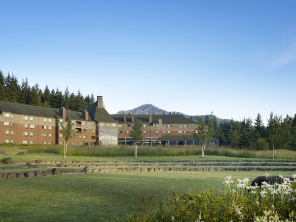 A large brick hotel or lodge set against a forested backdrop with a distant mountain, a manicured lawn, and white flowers in the foreground.