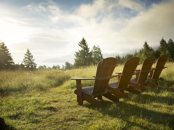 A serene lakeside meadow with four wooden Adirondack chairs facing the horizon as the sun sets behind trees.