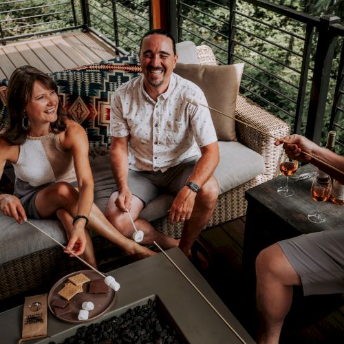 Three people are seated outdoors, roasting marshmallows and smiling, with a table holding s'mores ingredients and drinks nearby.