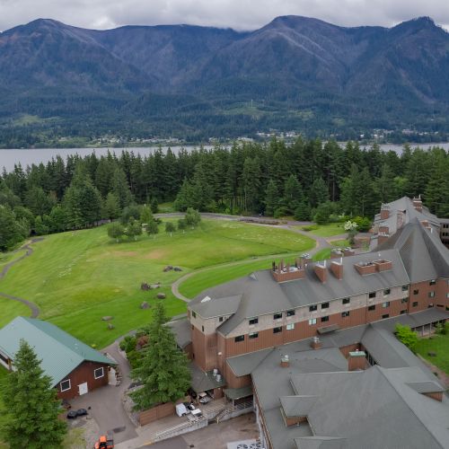 Aerial view of a large lodge surrounded by lush greenery, forest, and a lake with mountains in the background under a cloudy sky.