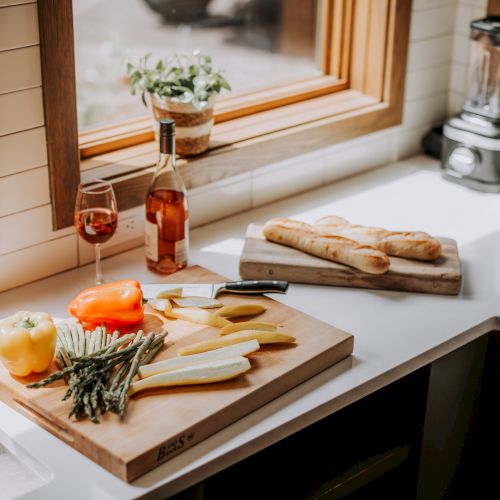 A kitchen counter with chopped vegetables, a bottle of wine, a glass, a loaf of bread, a toaster, and a blender near a window.
