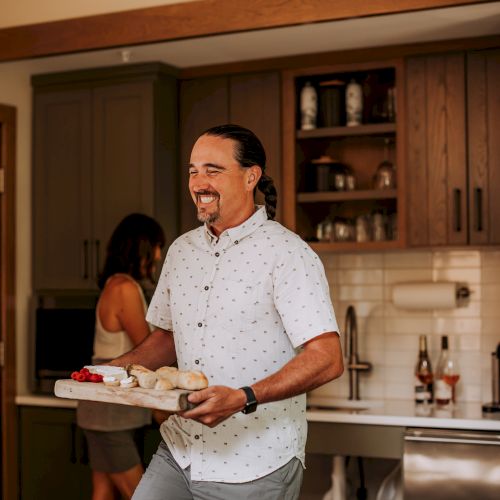 A man in a kitchen carries a tray of food while smiling; a woman is in the background by the sink.