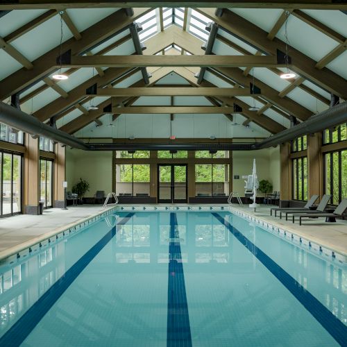 An indoor swimming pool with high ceilings, wooden beams, lounge chairs, and large windows offering natural light, creating a relaxing atmosphere.