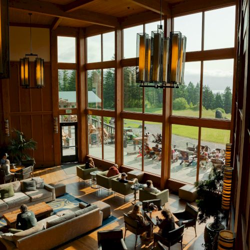 A spacious lodge interior with large windows, cozy seating, and people relaxing, offering views of a green landscape and distant water.