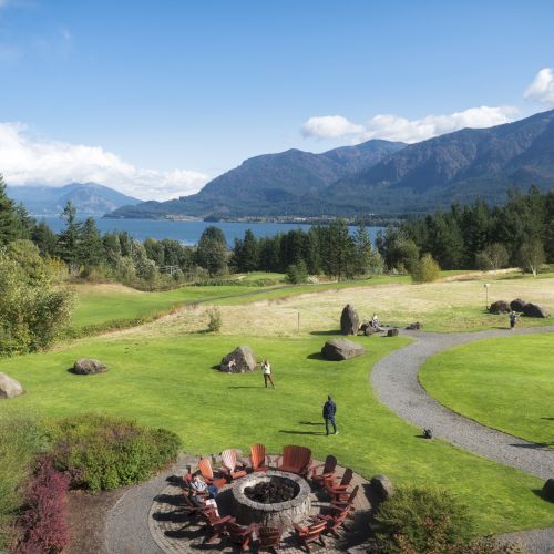 A scenic landscape with mountains, a lake, and people walking near a firepit in a grassy area surrounded by trees.