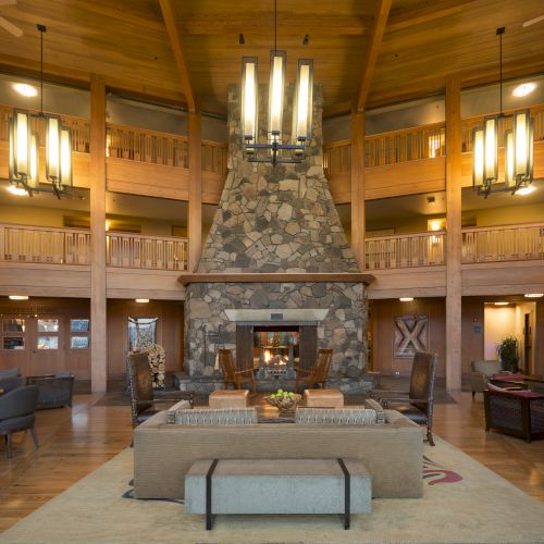 Spacious lodge interior with a central stone fireplace, wooden beams, chandeliers, and cozy seating area, creating a warm atmosphere.