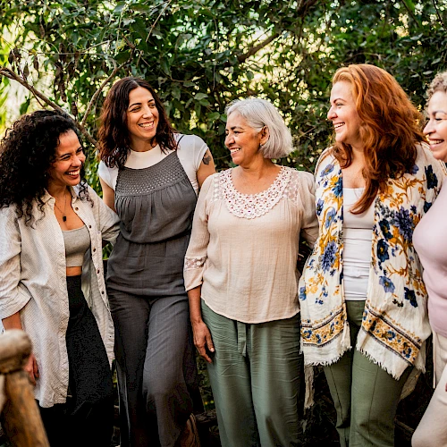 A group of five diverse women of different ages chat happily outdoors on a wooden railing path among green trees, sharing smiles.