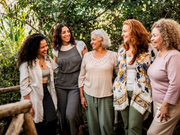 Five women of different ages stand together outdoors, smiling and chatting on a wooden railing path among greenery, enjoying each other&rsquo;s company.