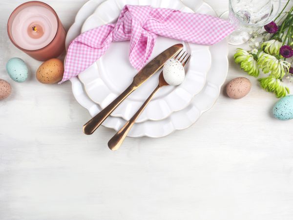 A table setting with a white plate, wooden cutlery, a pink napkin bow, pastel eggs, a pink candle, and spring flowers in a bright, airy layout.