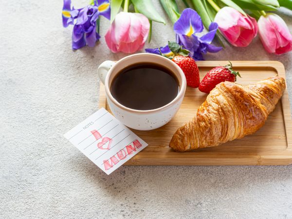 A cup of black coffee with a croissant on a wooden tray, strawberries, flowers, and a small card on a light table.