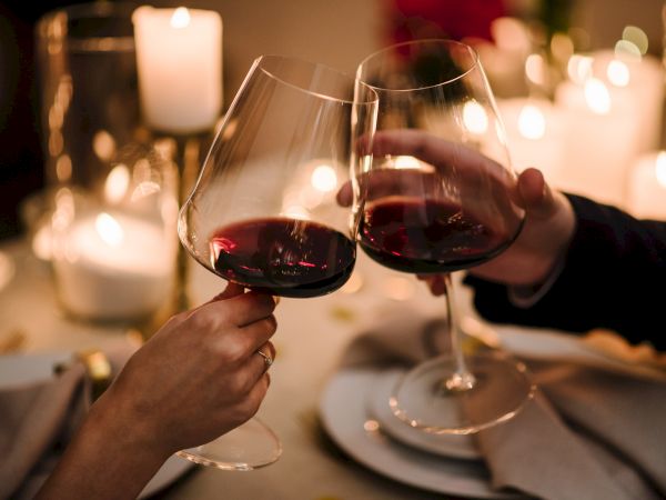 Two people clink wine glasses at a candlelit dinner, toasting in a warm, intimate setting.