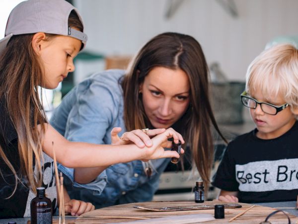 Three people&mdash;a woman and two kids&mdash;are at a table doing a hands-on nail or craft project with bottles and tools; they&rsquo;re focused and collaborative.