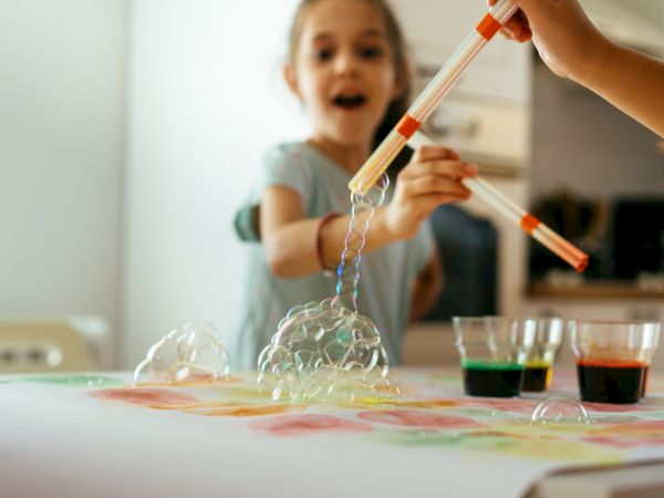 Children are playing with a bubbly science experiment at a table, using droppers to create foam above tinted cups.