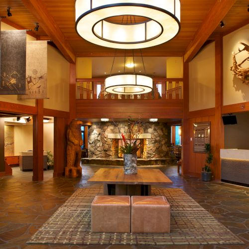 The image shows a spacious, warmly lit hotel lobby with modern decor, wooden accents, and a seating area arranged on a patterned rug.
