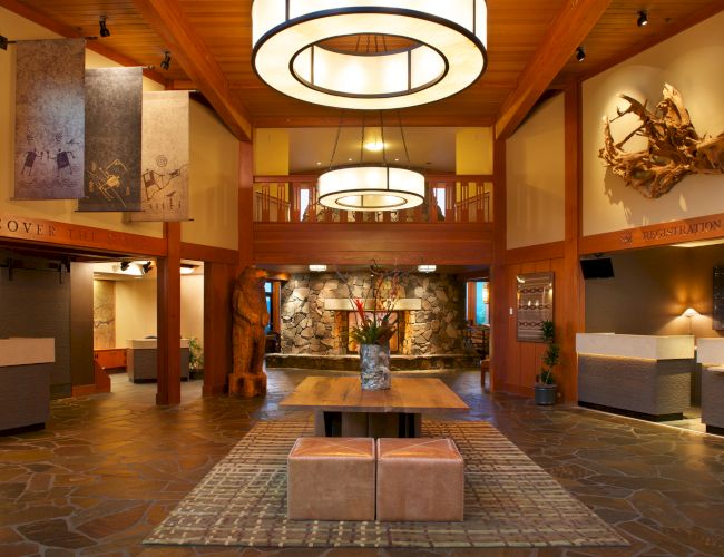 The image shows a spacious, warmly lit hotel lobby with modern decor, wooden accents, and a seating area arranged on a patterned rug.