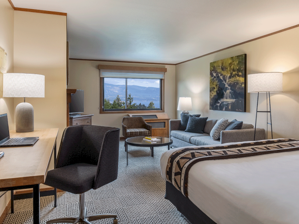 A hotel room with a bed, desk, chair, sofa, and lamps, plus a window view.