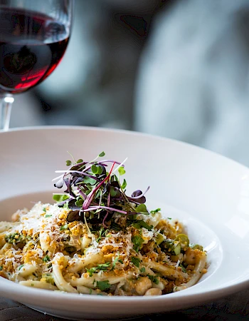 A bowl of pasta with greens and microgreens, topped with parmesan, beside a glass of red wine.