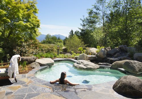 Person relaxing in a hot tub surrounded by rocks and greenery, with mountains in the distance, on a sunny day.