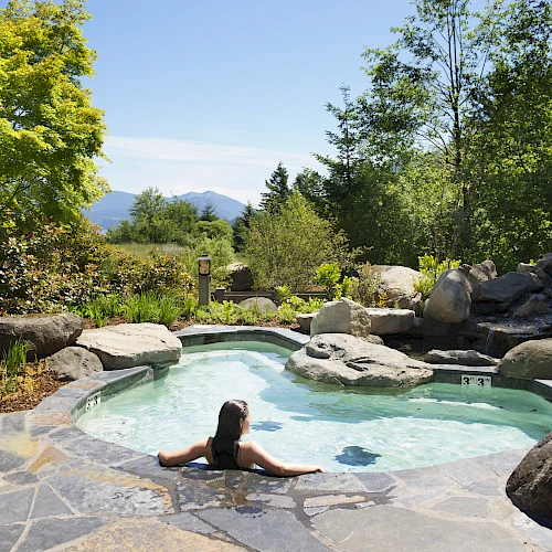 Person relaxing in a hot tub surrounded by rocks and greenery, with mountains in the distance, on a sunny day.