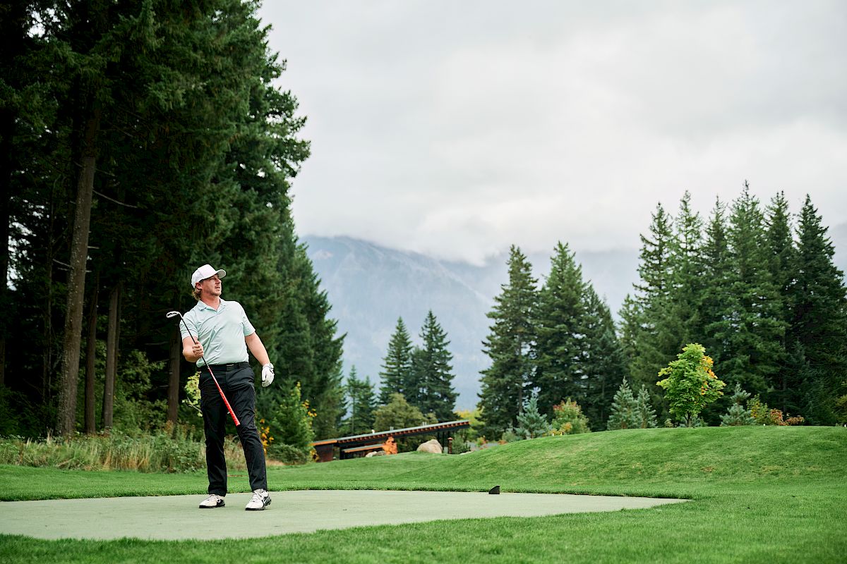 A golfer in a light shirt and cap stands on a tee, aiming with a club on a lush, park-like course with trees and mountains in the background.
