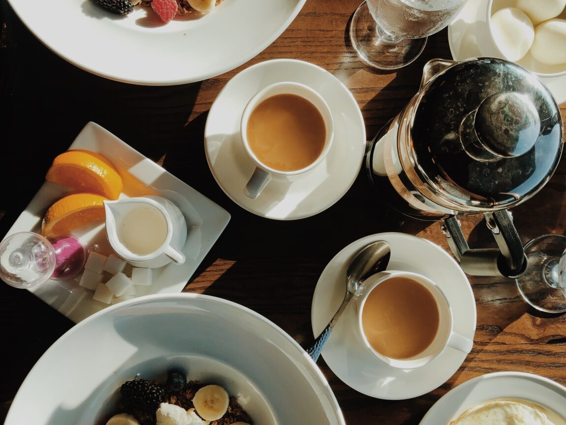 A table with two bowls of yoghurt topped with berries and banana slices, plus coffee, iced water, cream, lemon slices, and a small plate of fruit and jelly, tasty and wholesome.