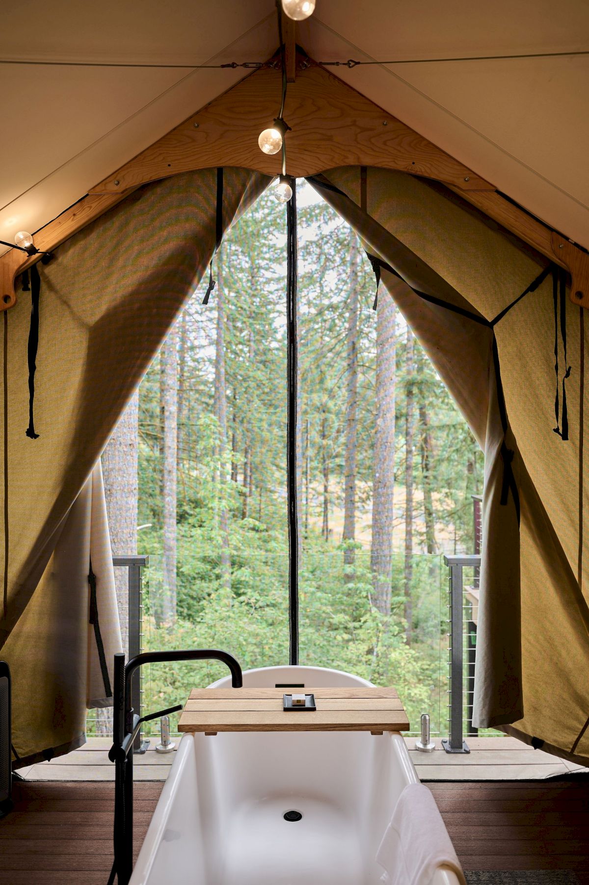Inside a tent with a view of a forest, a freestanding bathtub sits on a wooden deck, a faucet on the side and string lights above.