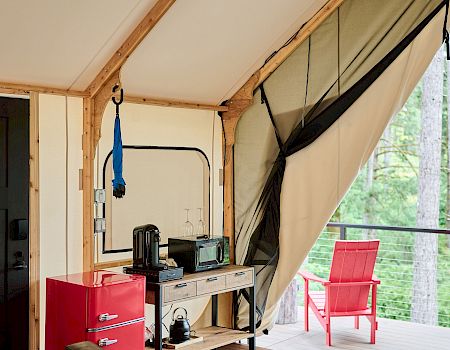 Cozy cabin interior with a desk, red mini-fridge, coffee maker, seating area, string lights, and a tented wall opening to a balcony with a red chair.