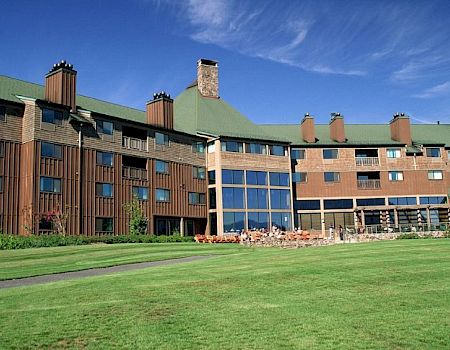 A large wooden-and-brick hotel or lodge-like building with green roofs, many windows, and a wide lawn in front, under a blue sky.