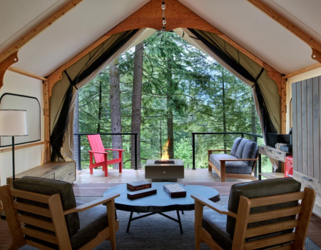 A cozy cabin loft with wood furniture, a central coffee table, a lit fireplace, and a view of lush trees through a veranda.