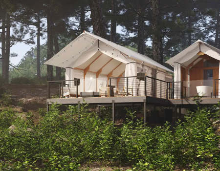 Two white A-frame cabins on a deck among pine trees, with outdoor seating and a railing, overlooking greenery.