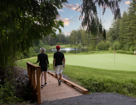 Two people walk a wooden bridge toward a golf green by a pond, surrounded by trees on a sunny day.