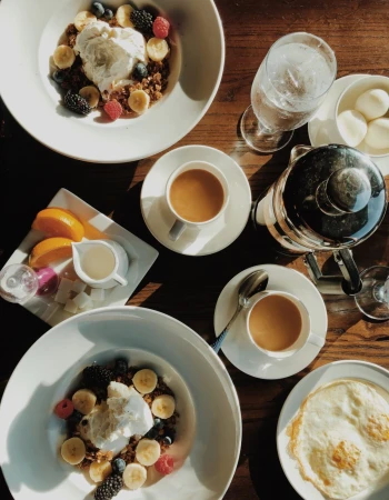 A top-down breakfast scene with bowls of yogurt topped with berries and banana slices, a fruit platter, espresso, cream, and a glass of water.