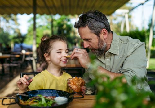 A man feeds a smiling girl at an outdoor cafe, sharing a sweet moment over lunch together.