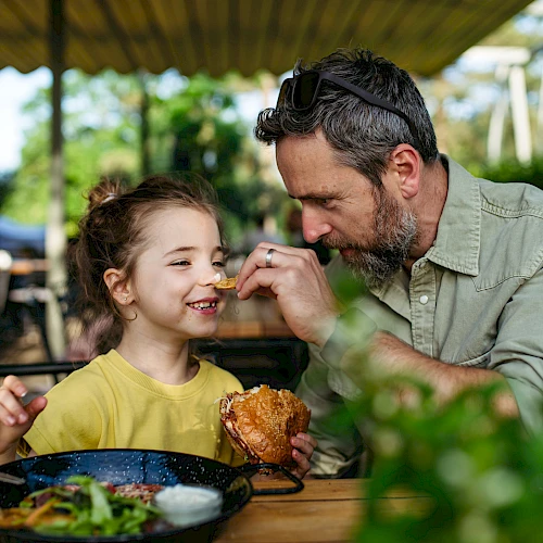 A man feeds a smiling girl at an outdoor cafe, sharing a sweet moment over lunch together.