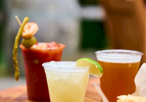 A wooden table with assorted drinks (pale yellow, amber, and red) and a basket of snacks; lime wedge garnish on one drink, cocktail sticks visible.