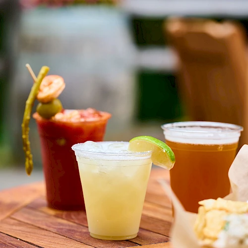 A wooden table with assorted drinks (pale yellow, amber, and red) and a basket of snacks; lime wedge garnish on one drink, cocktail sticks visible.