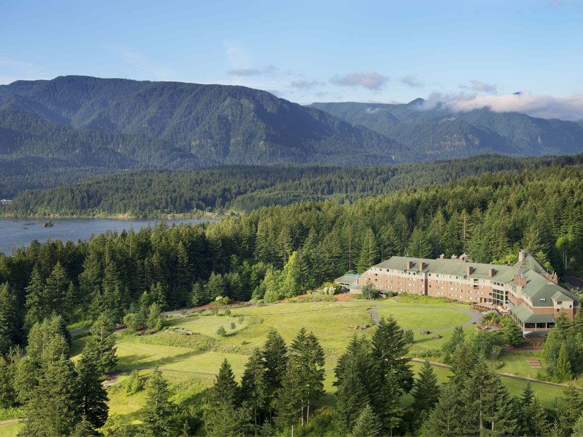 A hillside lodge surrounded by dense evergreen forests, with a long building complex near a grassy yard, and distant mountains under a clear sky.