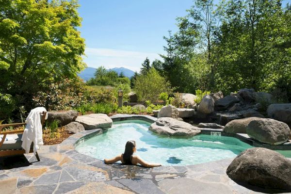 A person relaxes in a small, naturalistic pool surrounded by stones and greenery with mountains in the background on a sunny day.