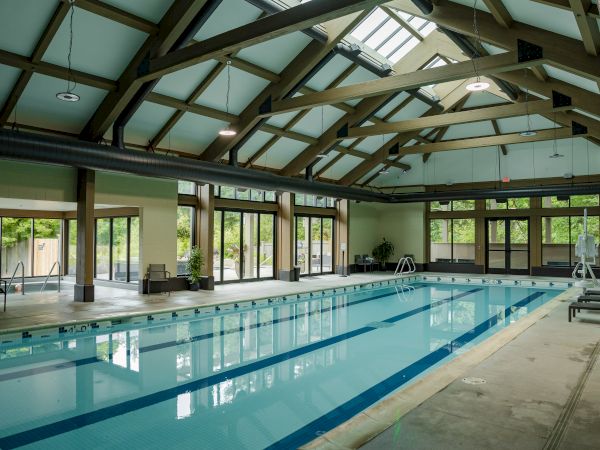 An indoor swimming pool with a high ceiling, large windows, and seating areas, surrounded by wooden beams and natural lighting.