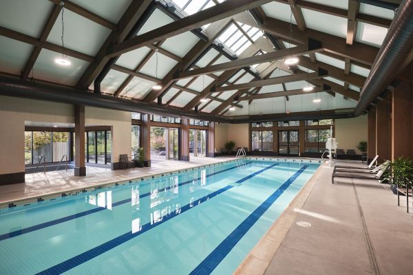 An indoor swimming pool with wooden beams, skylights, lounge chairs, and glass doors leading to an outdoor area.