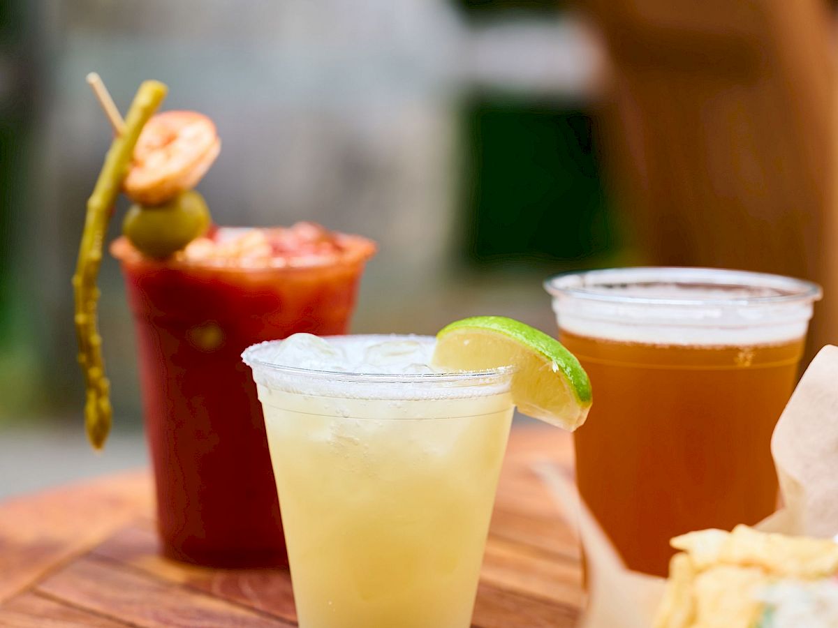A sunny outdoor scene with three drinks (one yellow with lime, two amber/fruit drinks) and a sandwich on a wooden table, blurred background.