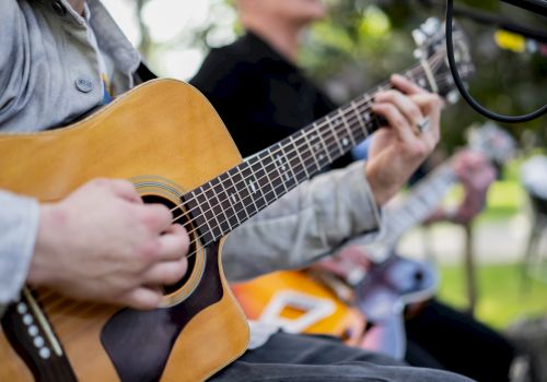 A person plays an acoustic guitar outdoors, focusing on strumming near the soundhole, with another guitarist visible in the blurred background.