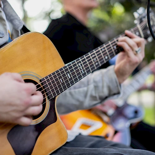 A person plays an acoustic guitar outdoors, focusing on strumming near the soundhole, with another guitarist visible in the blurred background.