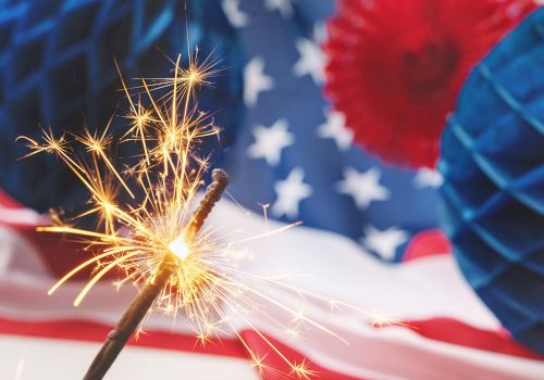 A sparkler burns brightly in the foreground, with an American flag backdrop and blue party decorations in celebration.