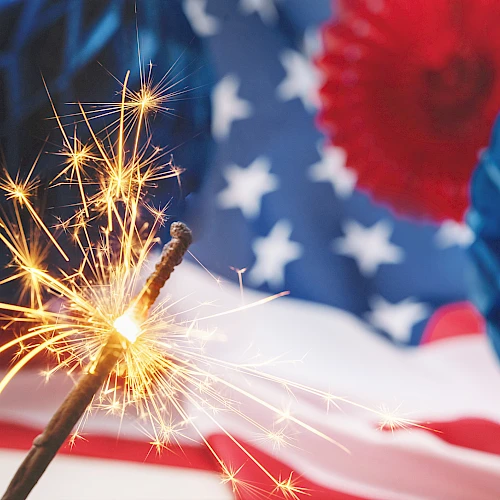 A sparkler burns brightly in the foreground, with an American flag backdrop and blue party decorations in celebration.