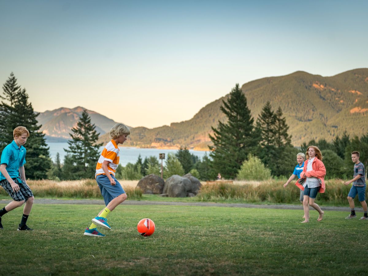 Kids playing soccer on a grassy field with mountains, trees, and a lake in the background.
